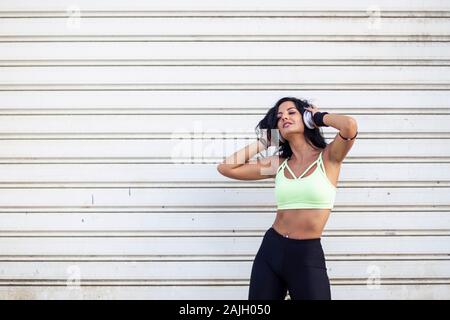 Fitness Mädchen mit langen dunklen Haaren stand vor der Tür mit Kopfhörern über den Kopf Stockfoto