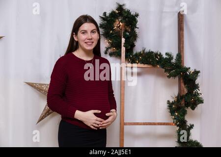 Schöne smiley schwangere Mädchen, die auf dem Hintergrund der Weihnachtsdekoration. Weihnachten oder Neujahr Atmosphäre. Stockfoto