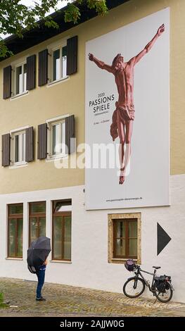 Frau mit Regenschirm steht im Regen und blickt auf die prächtige Wandwerbung, die das bevorstehende Passionsspiel vor der Oberammergauer Kasse darstellt Stockfoto