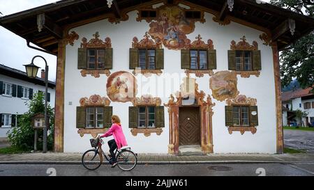 Rot geköpft Radfahrerinnen reitet an einer erstaunlichen Hausfassade vorbei mit im bayerischen Stil gemalten Freskos namens Lüftlmalerei. Stockfoto