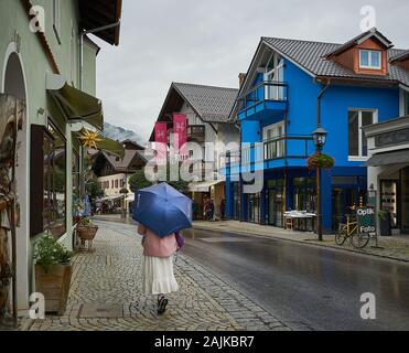 Modische Frau mit Regenschirm schlendern durch die malerischen Dorfstraßen von Oberammergau in Bayern, die für ihr 10-jähriges Passionsspiel bekannt sind. Stockfoto