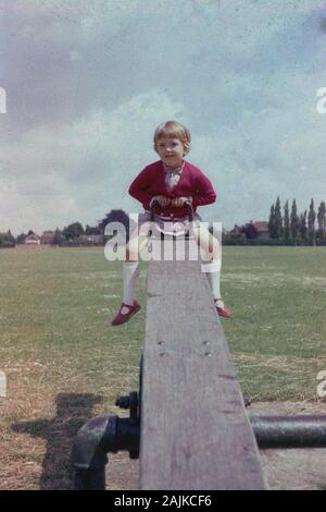 1960er Jahre, historisches, junges Mädchen in einem roten Oberteil und Sandalen, die auf einer Holzseesaw draußen auf einem Parkspielplatz sitzen, England, Großbritannien. Stockfoto