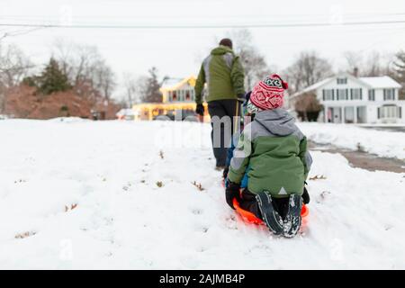 Rückansicht des Vaters, der Kinder auf einem orangen Schlitten durch den Schnee zieht Stockfoto