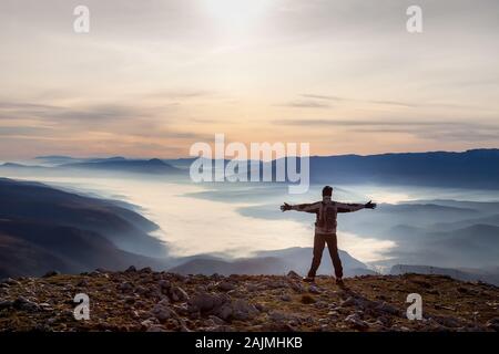 Bergwanderer, die am Rande des Berggipfels mit Händen in der Luft stehen und ein Tal mit dichtem Nebel bei farbenfrohem Sonnenaufgang betrachten Stockfoto