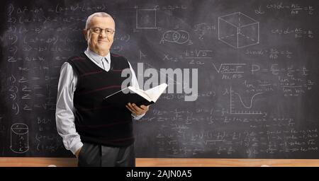 Ältere männliche Lehrer mit Buch und stand vor einer Tafel mit mathematischen Formeln Stockfoto