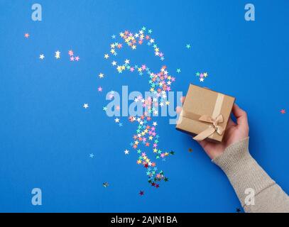 Weibliche Hand in einem goldenen Pullover halten geschlossen Square blue box mit einem Bogen auf einem blauen Klassischer Hintergrund, Ansicht von oben, festliche Kulisse Stockfoto