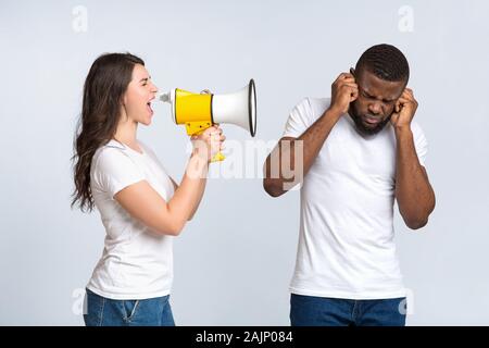 Gereizte junge Frau schreiend mit Megafon an ihren Mann, dass die Schließung Ohren, weil der Lärm, Licht studio Hintergrund mit freier Speicherplatz Stockfoto