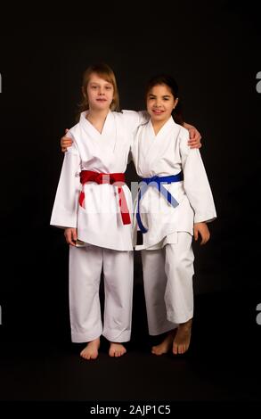 Zwei kleine Karate Mädchen Freunde in Weiß Kimonos, eine in Rot und die andere blau Wettbewerb Riemen umarmen vor einem dunklen Hintergrund Stockfoto