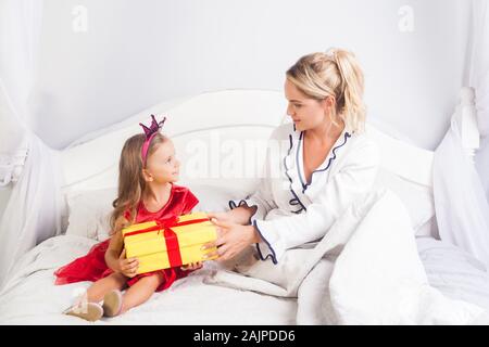 Junge Frau, die gegenwärtig zu wenig süße Mädchen in einem hellen Kleid und Krone auf dem Kopf, sitzt auf dem Bett, die Mutter, die Tochter zum Geburtstag, Gruß c Stockfoto