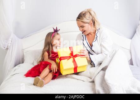 Junge Mutter Eröffnung Geschenkkarton mit überrascht, erstaunt, Gesicht, Sitzung mit niedlichen kleinen Tochter auf dem Bett, Kind, Frau, alles Gute zum Geburtstag, Gruß Mama o Stockfoto