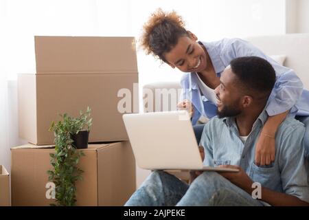 Aufgeregt Afro Ehegatten mit Laptop Computer sitzen auf dem Sofa unter Kisten ausgepackt im neuen Haus. Freier Speicherplatz, selektiven Fokus Stockfoto