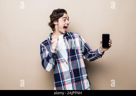 Porträt einer aufgeregten Jungen Mann stehend über beigen Hintergrund isoliert, holding Handy Stockfoto