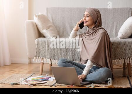 Arbeitsmöglichkeiten für muslimische Frauen Konzept. Lächelnd Arabische Mädchen im Hijab Gespräch am Handy und Laptop beim Sitzen auf dem Boden zu Hause. Stockfoto