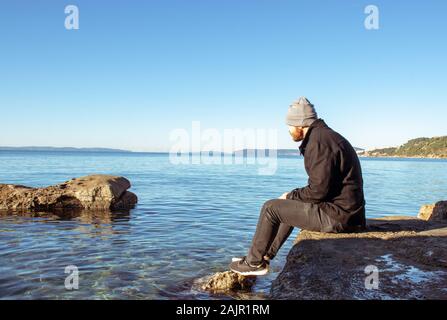 Bärtiger Mann mit Mütze und eine schwarze Hose und Jacke sitzt auf einem Felsen an der Küste der Adria. Blaues Wasser und Inseln in der Ferne. Idyllische Stockfoto
