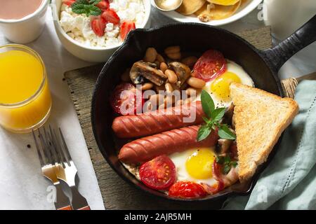 Keto Frühstück: Spiegeleier, Würstchen, Bohnen, Speck, Pilzen, gegrillte Toast, Orangensaft, Quark, Pfannkuchen und Kakao auf einem Tisch aus Stein Stockfoto