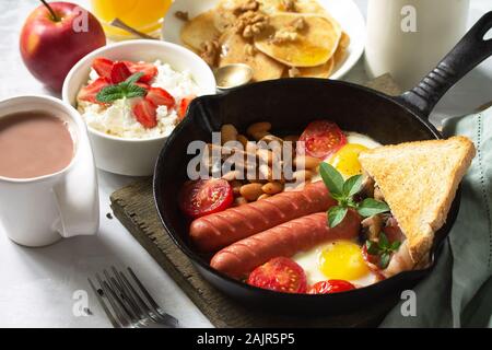 Keto Frühstück: Spiegeleier, Würstchen, Bohnen, Speck, Pilzen, gegrillte Toast, Orangensaft, Quark, Pfannkuchen und Kakao auf einem Tisch aus Stein Stockfoto