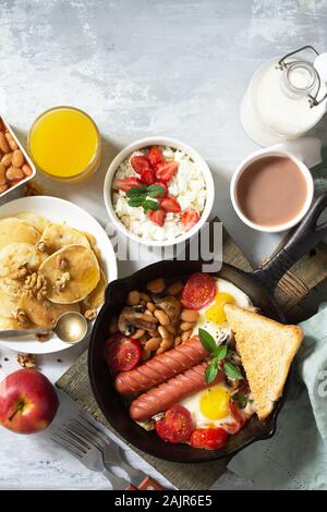 Keto Frühstück: Spiegeleier, Würstchen, Bohnen, Speck, Pilzen, gegrillte Toast, Orangensaft, Quark, Pfannkuchen und Kakao auf einem Tisch aus Stein Stockfoto