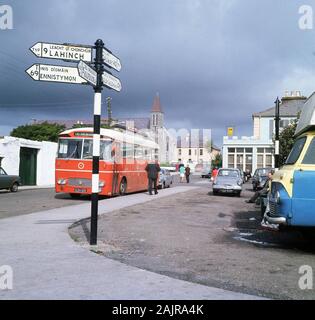 In den 1960er Jahren parkte ein roter öffentlicher Bus am Straßenrand in der Kurstadt Lisdoonvarna, County Clare, Irland, und zeigte in der Ferne den Turmvorbau der viktorianischen gotischen Wiederauferstehung der Römisch-katholischen Kirche. In dieser Epoche berühmt für sein Musikfestival, ist es jetzt eine "Matchmatching"-Veranstaltung, eine der größten Europas, die die größte Touristenattraktion der Stadt ist. Stockfoto