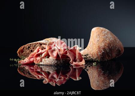 Prosciutto mit Ciabatta und Thymian auf schwarzem reflektierenden Hintergrund. Stockfoto