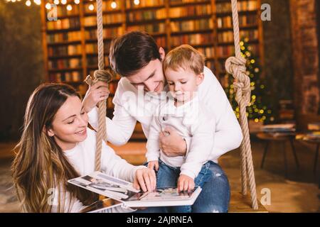 Familie mit Baby Entspannen auf Startseite Hollywoodschaukel in gemütlichen dunklen Wohnzimmer am Heiligabend. Familie Spaß auf Wippe. Winter Abend mit Familie und Stockfoto