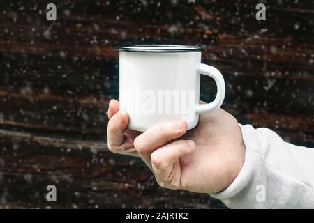 Nahaufnahme von Mann reisende Hand metall Becher mit heißen Getränk mit unscharfen fallenden Schnee. Outdoor Tee, Kaffee entspannen. Mockup von weißen Emaille Tasse, Stockfoto