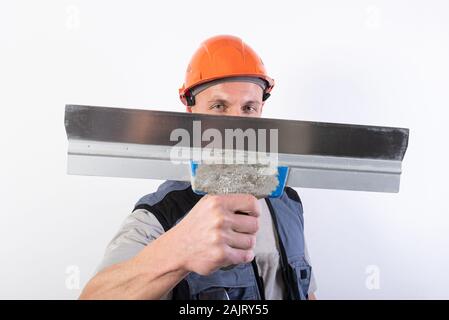 Die Builder verbirgt sein Gesicht hinter einem Spachtel. In Arbeitskleidung und harten Hut. Auf einem hellgrauen Hintergrund. Stockfoto