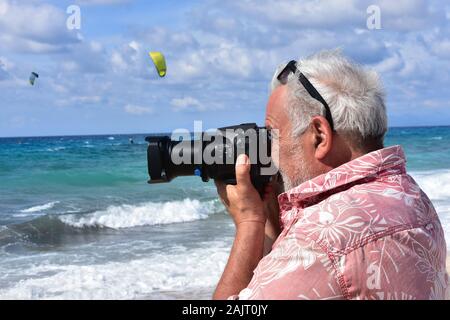 Älterer Mann am Strand shooting Video mit seiner Kamera; unscharf Kiter Surfen im blauen Meer im Hintergrund Stockfoto