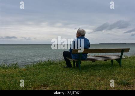 Ältere Mann sitzt auf einem alten Holzbank, über die in einem bewölkten Tag sehen, betrachten die nostalgischen Blick aufs Meer Stockfoto