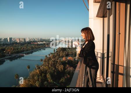 Junge Frau lächelnd und Trinken einige Getränke- und Blick vom Balkon. Stockfoto