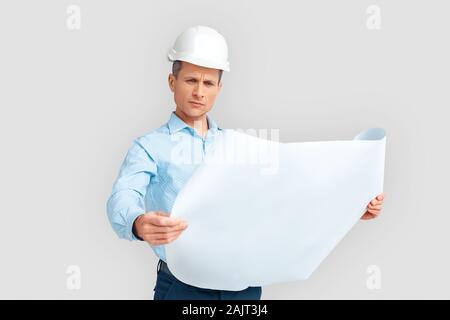Konstruktion. Reifer Mann in hardhat Ständigen isoliert auf Weiss mit blueprint Studium betreffenden Stockfoto