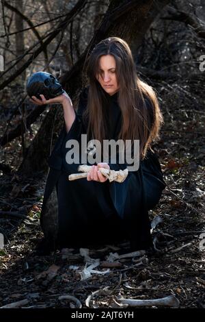 Frau in schwarzen Roben mit Haufen von grossen wilden tierischen Knochen in der Mitte des Waldes. Magie und Hexerei Konzept. Stockfoto