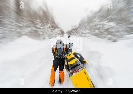 Ein Mann, ein Athlet in einem warmen Sportanzug Fahrten mit einem Schneemobil auf einer verschneiten Straße vor dem Hintergrund einer Winter wald landschaft Stockfoto