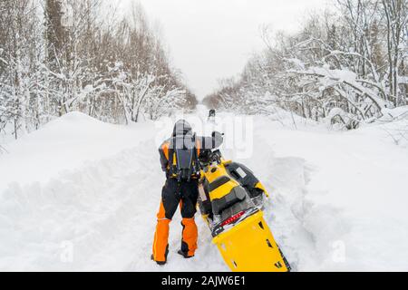 Ein Mann, ein Athlet in einem warmen Sportanzug steht und schiebt ein Schneemobil auf einer verschneiten Straße vor dem Hintergrund einer Winter wald landschaft Stockfoto