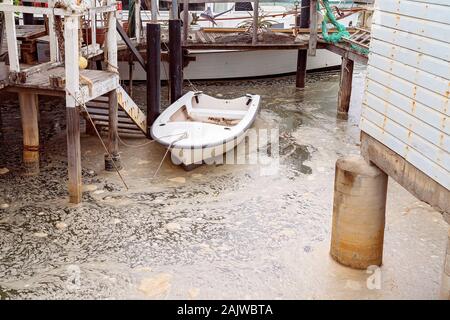 Eine kleine weiße Zeile boot gebunden bis zu einem Post in wirbelnden schaumig schmutziges Wasser in einem alten Fachwerkhaus Bootsanleger mit Müll beladen Stockfoto