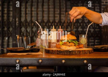 Hand, die Messer mit Burger auf Holzplatte mit Sodawasser. Stockfoto