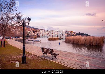 Stimmungsvoller Blick auf die Stadt Kastoria und den See Orestiada bei Sonnenuntergang. Die Promanade mit Bank und Straßenbeleuchtung im Vordergrund. Stockfoto