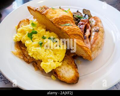 Ein ganztägiges Frühstück/Brunch combo Mahlzeit Teller mit Rührei, Croissants, Wurst, Speck und Pilzen auf eine weisse Platte bei einer Western casual Resta Stockfoto