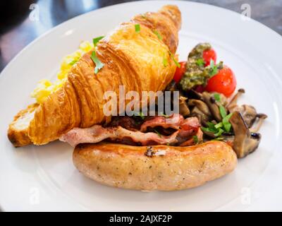 Ein ganztägiges Frühstück/Brunch combo Mahlzeit Teller mit Rührei, Croissants, Wurst, Speck und Pilzen auf eine weisse Platte bei einer Western casual Resta Stockfoto