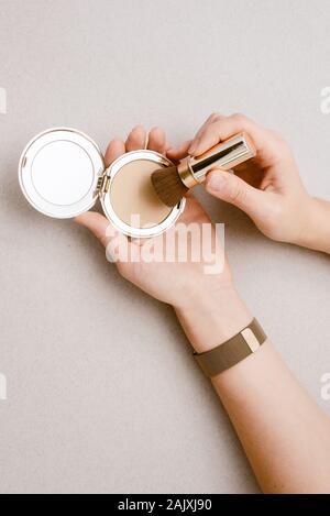 Compact Powder mit einem Spiegel in der Hand eines jungen Mädchens, schließen. Gedrückt klangliche Basis in teure goldene Verpackung auf einem beigen Hintergrund. Das Mädchen Colle Stockfoto