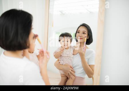 Lustige Familie zu Hause. Mutter und Kind Mädchen tun Sie Ihr Make-up und Spaß haben in der Nähe von Spiegel. Baby Mädchen erforscht die Mutter Kosmetik zu Hause Stockfoto