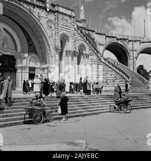 In den 1950er Jahren waren Besucher am Eingang zur Rosary Basilica, Lourdes, Frankreich, einer katholischen Kirche am "Heiligtum unserer Lieben Frau von Lourdes" oder der Domäne, einem Erdgebiet rund um den katholischen Schrein (Grotte), zu sehen. Ein Pilgerort für viele des christlichen Glaubens, vor allem für kranke und unfeste, wie man auf dem Bild der Menschen in den ungültigen Kutschen der Epoche erkennen kann. Stockfoto