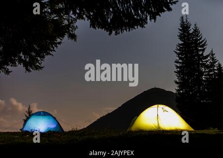 Zwei hell leuchtende, touristische Zelte auf der grünen Wiese Wald Lichtung auf dunklen Berg und dem klaren, blauen Sternenhimmel Kopie Raum Hintergrund. Tourismus, Nacht campin Stockfoto