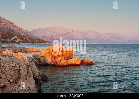 Felsige Küste des kroatischen Meeres, Meer und Felsen im Vordergrund, Klippen und Berge im Hintergrund, beleuchtet von der untergehenden Sonne Stockfoto