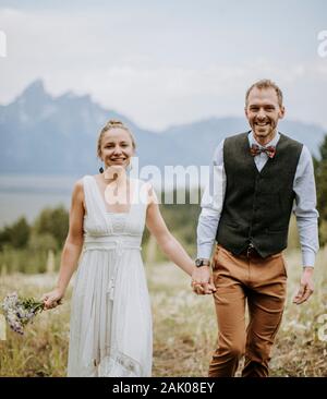 Glücklich lächelnd frisch vermählte Braut und Bräutigam gehen durch Feld in Tetons Stockfoto