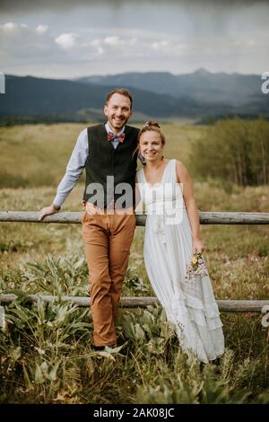 Lächelnde glückliche Braut und Bräutigam lehnen sich gegen Zaun im Feld in Wyoming Stockfoto