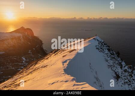 Weibliche Wanderer steht allein auf verschneiten Berggipfel mit Sonne im Hintergrund, Flakstadøy, Lofoten, Norwegen Stockfoto