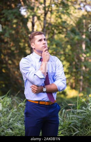 Junger Mann im Anzug steht draußen, Hand auf Kinn, tief im Gedanken. Stockfoto