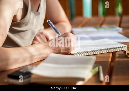 Nahaufnahme einer Frauenhand mit einem Stift, schreibt in ein Notizbuch, legt ein Buch auf den Tisch, Handy, Bleistifte, im Freien auf einer Terrasse auf einem sonnigen d Stockfoto