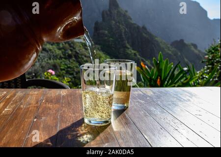 Man gießt Weißwein aus Ton Kanne in Glas auf der Terrasse mit Blick auf die grüne Landschaft der kleinen Bergdorf Masca auf Teneriffa, Spanien im sonnigen d Stockfoto