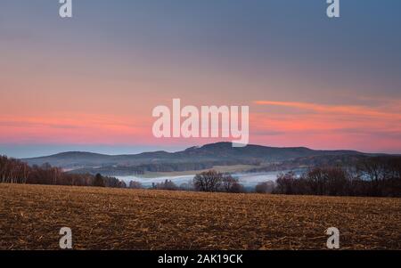 Landschaft bei Sonnenuntergang, im Vorderfeld und Bäume, im Tal weißer Nebel, im Hintergrund Wald und Berge, roter und blauer Himmel Stockfoto